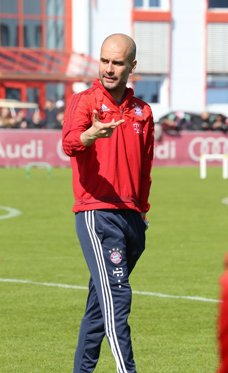 Pep Guardiola berjaket merah FC Bayern München memberi instruksi dengan gerakan tangan di lapangan latihan