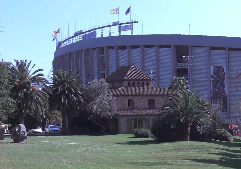 Bangunan batu La Masia dikelilingi pohon palem dengan tribun Camp Nou di belakangnya