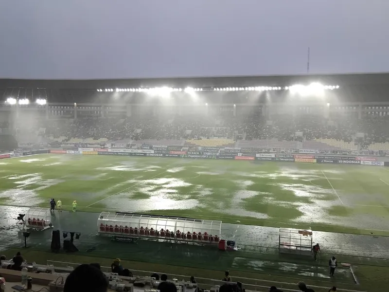 Lapangan Stadion Manahan tergenang air hujan di bawah sorot lampu stadion, bangku cadangan di tepi lapangan