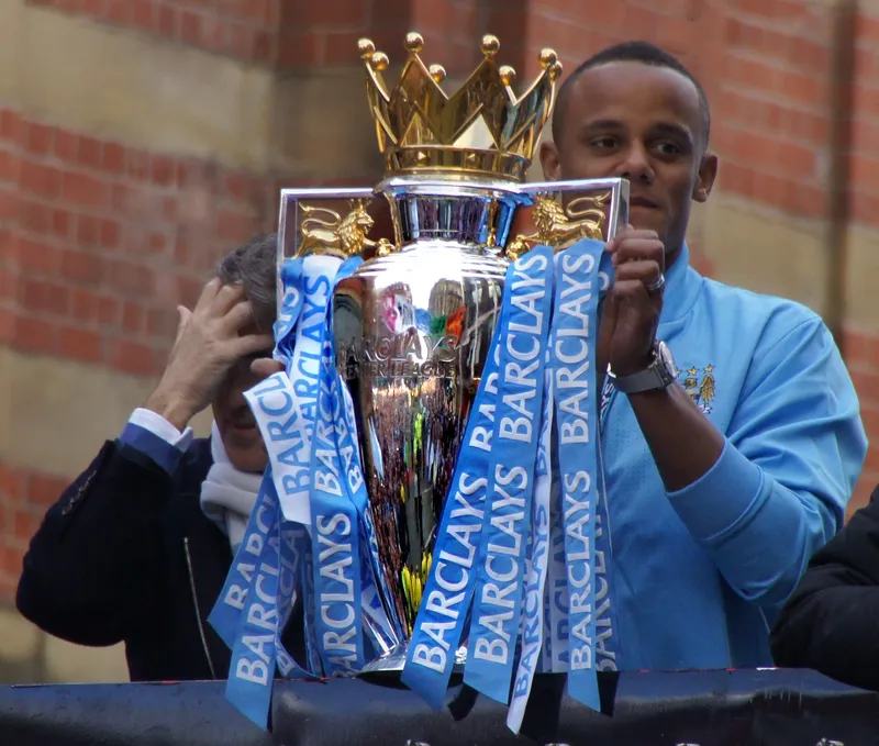 Vincent Kompany berjaket biru muda memegang trofi Premier League berpita Barclays