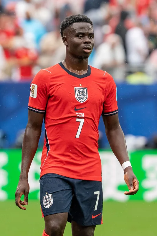 Bukayo Saka berdiri dengan seragam merah Inggris nomor 7.