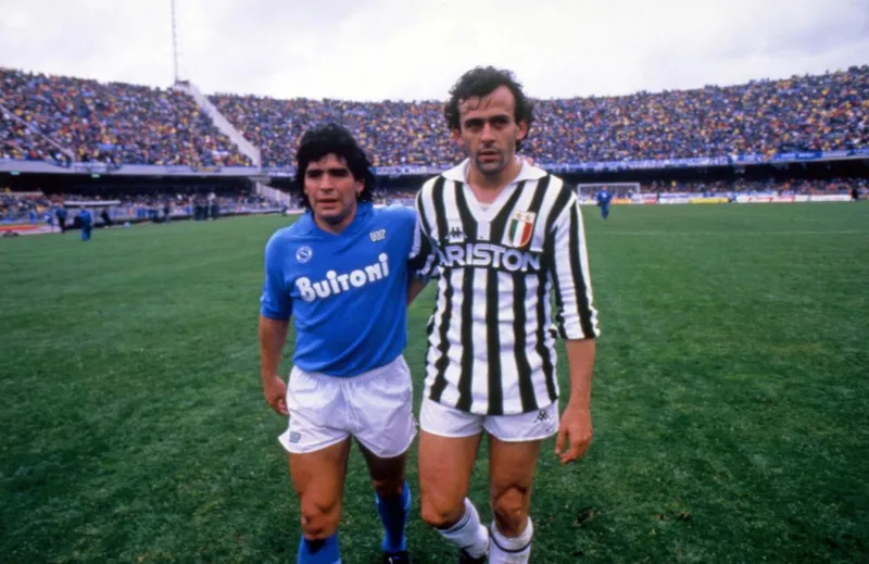 Diego Maradona berseragam biru Napoli berdiri di samping Michel Platini berseragam garis hitam putih Juventus di lapangan stadion
