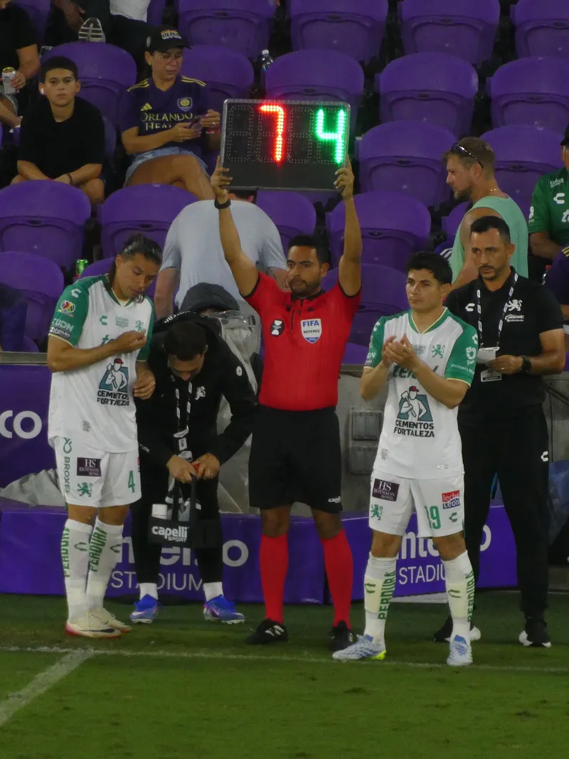Ofisial keempat berbaju merah mengangkat papan elektronik bertuliskan 7 dan 4 di antara dua pemain Club Leon