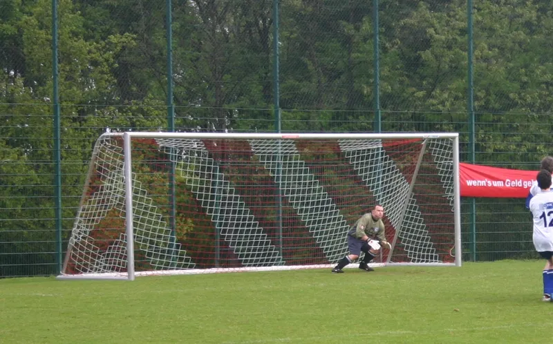 Kiper berseragam hijau bersiap di depan gawang sepakbola ukuran penuh berjaring putih merah